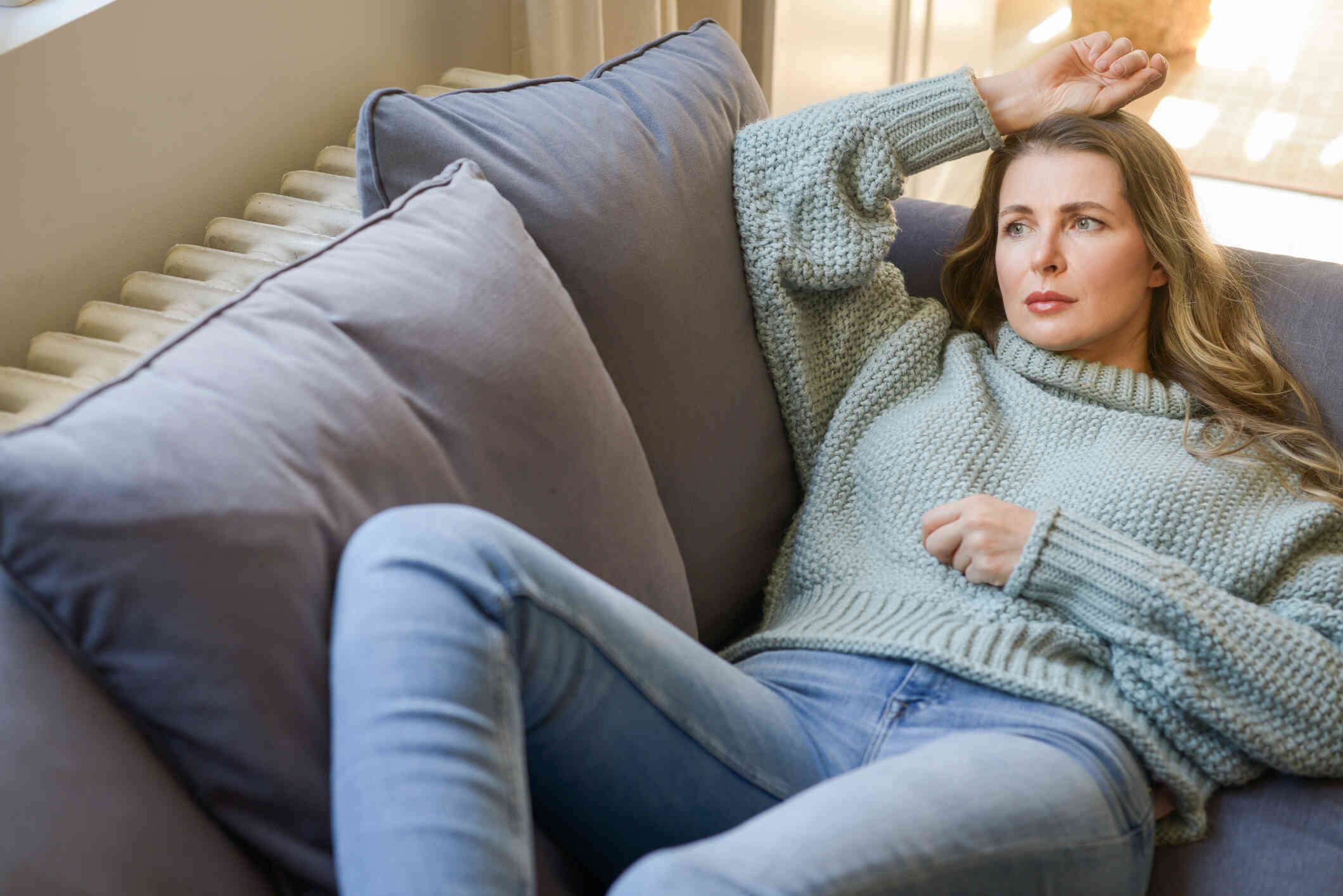 A woman in a green sweater lays on her back on the couch with her arm rested across her forehead while she gazes off with a sad expression.